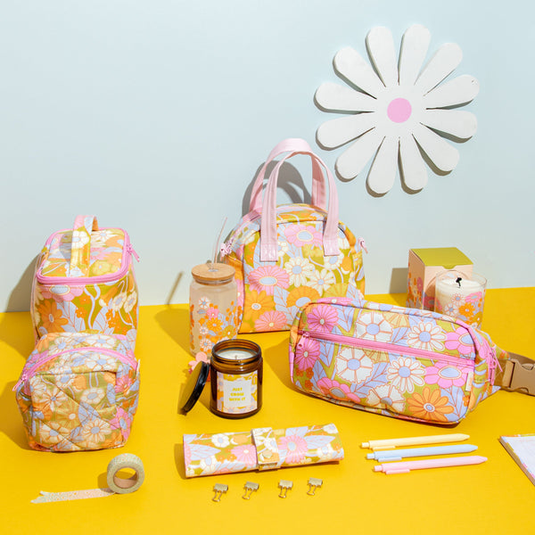 Flat lay of colorful vintage floral bags, pastel stationery, daisy-patterned glass can cup, cute candles, and cute desk accessories on a yellow background with a large paper daisy decor – perfect for spring vibes and cheerful organization.