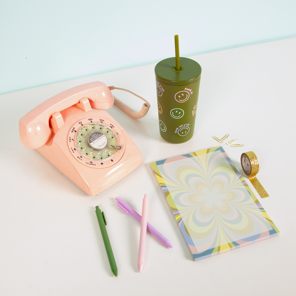 Retro-inspired flat lay featuring a pink rotary phone, green smiley face tumbler with straw, pastel swirl tearaway notepad, pink, purple, and green pens, gold glitter washi tape, and paper clips on a white desk with a light blue wall.