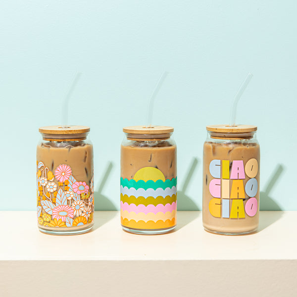 A trio of 3 clear can glass tumblers with a lid and clear straw. All three tumblers feature designs in multi 
pastel colors. All three tumblers are filled with iced coffee.