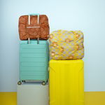 Light blue background with a nylon colorful smiley pattern stowaway tote attached to a baby blue suitcase. Next, there is a light violet and green checker pattern tote on top of a neon green suitcase.