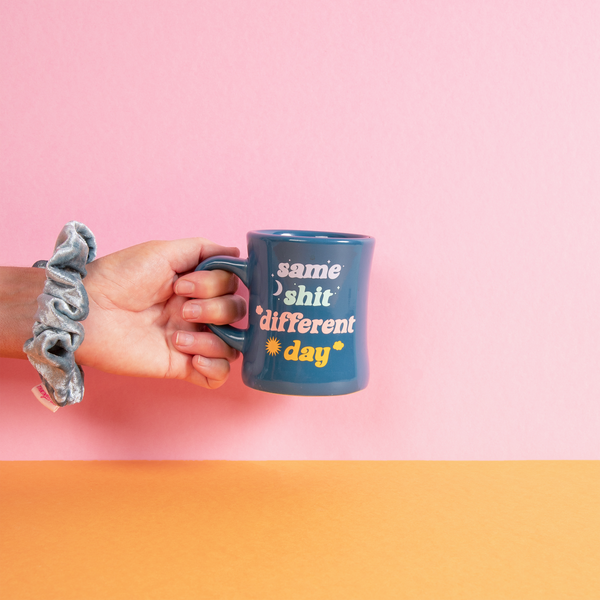 A hand holding a diner mug. The stylish mug features a dark teal background color with the saying "same shit different day" written in light blue, light green, pink, and orange. There is a moon and a sun to represent day and night.
