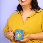 Smiling woman in a yellow shirt holding a blue diner coffee mug with bold text that says “Fuck Fuck Fuckity Fuck” and a cute smiley flower design, against a pastel purple background.