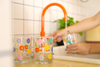 3 colorful retro daisy print rock glasses by a bright orange kitchen faucet, with a person washing a glass in a modern tiled kitchen setting. 
