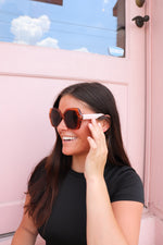 A smiling woman wearing oversized sunglasses with a transparent brown tinted frames and blush arms, posing in front of a pastel pink door with a clear blue sky reflection.