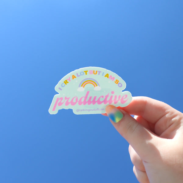 A hand holding a super honest sticker with the saying "I CRY A LOT BUT I AM SO PRODUCTIVE" The word productive is written in pink cursive while the rest of the words are written in alternating lavender, light blue, green, pink, and orange colors. There is a rainbow with clouds in the center of the sticker.