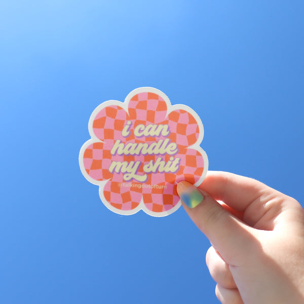 A hand holding a daisy shaped flower sticker. The sticker has a white outline and a super fun pink and orange checker pattern. The sticker has the saying "i can handle my shit" written in a cream color with purple outline written in cursive.