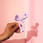 A hand holding a lilac claw clip in the shape of a bow. The lilac bow claw clip works on any length of hair and is a stylish accessory to any outfit.