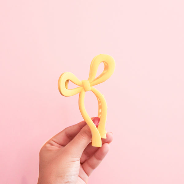 A hand holding a yellow claw clip in the shape of a bow. This yellow bow claw clip in a perfect way to add some cheerfullness to any outfit!!