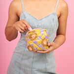 A cute girl in a denim romper standing against a blush pink background putting a lip gloss in a cute puffy pouch with a warped checkerboard pattern in citron and periwinkle with camel colored daisies scattered about.