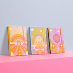 A set of 3 super cute colorful trippy, disco, and psychedelic notebooks with colorful elastic bands that hold them closed, standing in a row on a bright pink cube with a soft blue background.