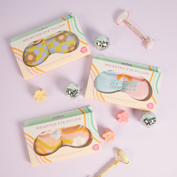 A flatly of 3 weighted eye masks in their cute Talkling Out Of Turn packaging on a lilac background in an assortment of patterns ranging from wavy checkered boards in citron and periwinkle with daisies scattered around to an ombre back ground with white daisies scattered around, and another that says "No More Mondays" with a rainbow in pink, orange, and yellow above it, with colorful floral hair clips and beautiful stone rollers, a true self care scene.