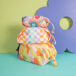 A colorful stack of three rectangular ripstop toiletry or makeup bags displayed against a playful, pastel backdrop. The top bag features a bold, multicolored retro floral pattern; the middle bag showcases a pastel checkered design with wavy squares in orange, lavender, mint, and yellow; and the bottom bag displays a groovy striped pattern in orange, green, pink, and yellow with scattered daisy flowers. Each bag has a matching zipper and side handle.