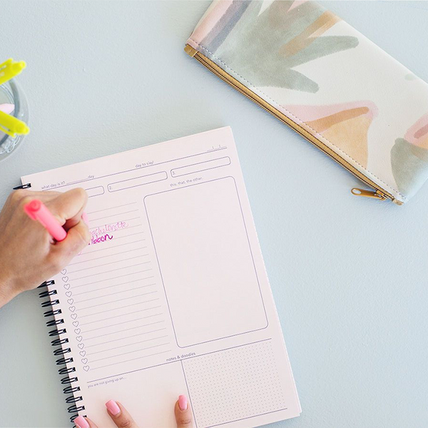 An above shot of a woman taking notes in a notebook with colorful pens and a small pencil pouch laying next to her.