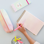 Hand writing in a taskpad with a cup of pens, a cute pencil pouch, and a taskpad nearby.