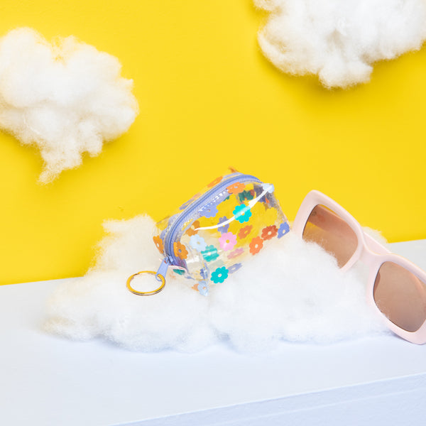 Colorful daisy pattern on a clear vinyl key pouch sitting on a cloud next to sunglasses. 