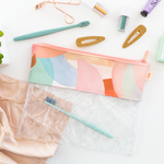 moonscape dollface pouch with half and crescent moon drawings in light blue, green, blush and peach sitting on a white table with hair clips toothbrush and make up accessories