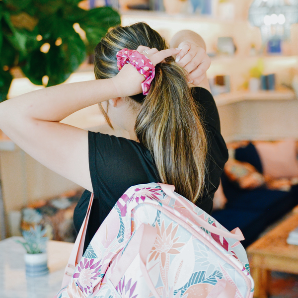 woman with brunette and blonde highlights putting a radiance collection Terrazzo speckled design scrunchie in a ponytail while wearing a floral nights backpack and black shirt.
