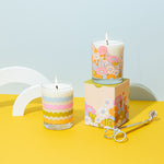 Rising scallops candle rockcs glass with gathering flowers pattern candle rocks glass on a mustard yellow surface with misc items. 