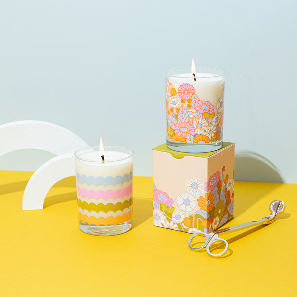 Rising scallops candle rockcs glass with gathering flowers pattern candle rocks glass on a mustard yellow surface with misc items. 