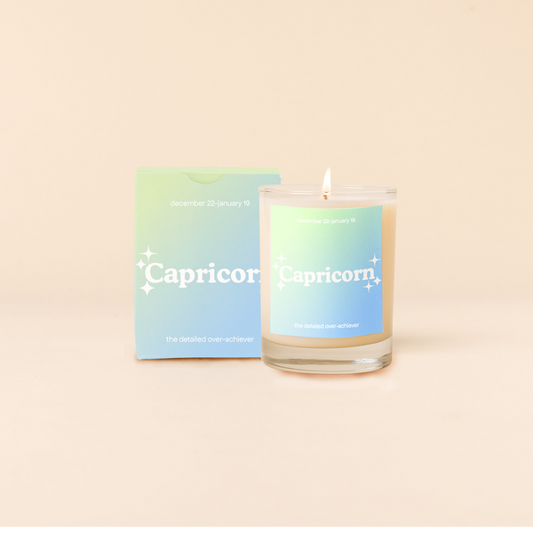 Candle rocks glass with sea foam green-to-light blue ombre decal and text that reads "Capricorn" with minimalist, white sparkle stars surrounding the text; "the detailed over-achiever" sits at the bottom of the decal. Box packaging with the same design sits behind glass.