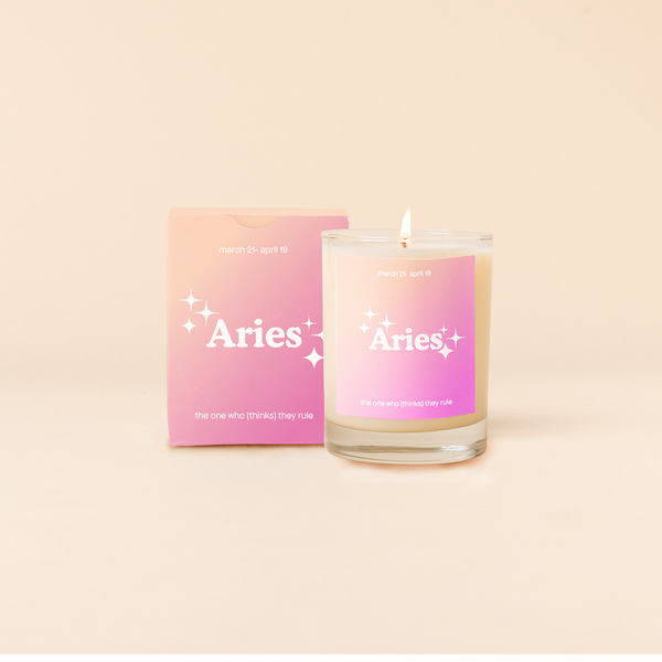 Candle rocks glass with cream-to-pink ombre decal and text that reads "Aries" with minimalist, white sparkle stars surrounding the text; "the one who thinks they rule" sits at the bottom of the decal. Box packaging with the same design sits behind glass.