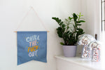 cornflower blue wall pendant saying chill the fuck out. On the side of the pendant there is a plant in a lilac plant holder, along with 2 metallic silver mushroom statues.