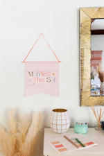Fairytail pink wall pendant saying manifest that shit. Hanging on the wall next to a gold rectangle mirror.