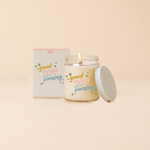 Glass jar candle reading "GOOD THINGS ARE COMING" in multi-color text surrounded by green stars. Box packaging with same design as candle. 