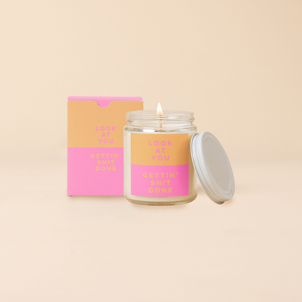 An 8 oz. candle w/ a lid and a split-colored decal (Orange and Pink) and the phrase "Look at you gettin' shit done" printed on.