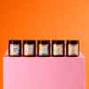 Amber candle jars with lids and delightful sayings lined up on a pink pedestal. 