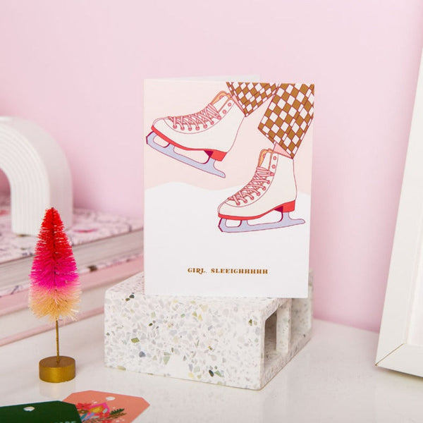 A white greeting card with Christmassy skates illustrated and "girl, sleighhhhh" written on the bottom