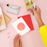 A white greeting card with graphic lettering "Wild Child". There is a red envelope, a gold tape dispenser and various desk items.