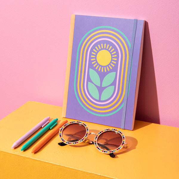 A periwinkle Journal with a sunflower on the cover leaning against a pink wall with sunglasses next to it. 