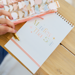 woman holding a gold jotter pen over a manifest that shit taskpad cover with stars gold wire binding and peach colored elastic closure strap