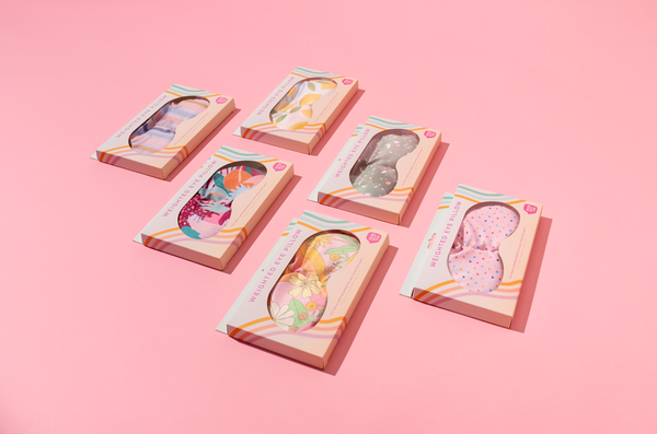 a pink weighted eye mask with assorted color tiny hearts in toots colorful packaging on a pink background.