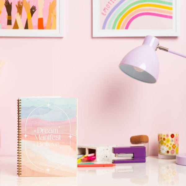 a griddy planner in water color pastels saying dream manifest believe surrounded by scattered stars with a coral elastic band to close it. 