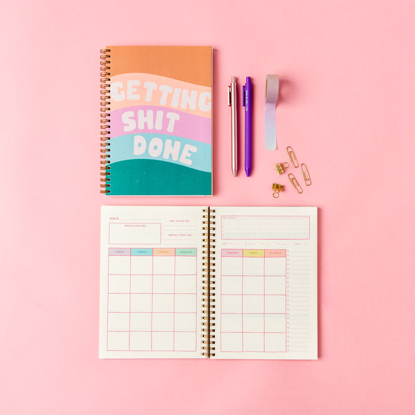 Perpetual Planner - Goal Getter Lite - Talking Out Of Turn undated "Getting Shit Done" planner closed and open. The open planner shows the undated monthly calendar while being surrounded by jotter pens, wash-tape, and paperclips on a pink background.