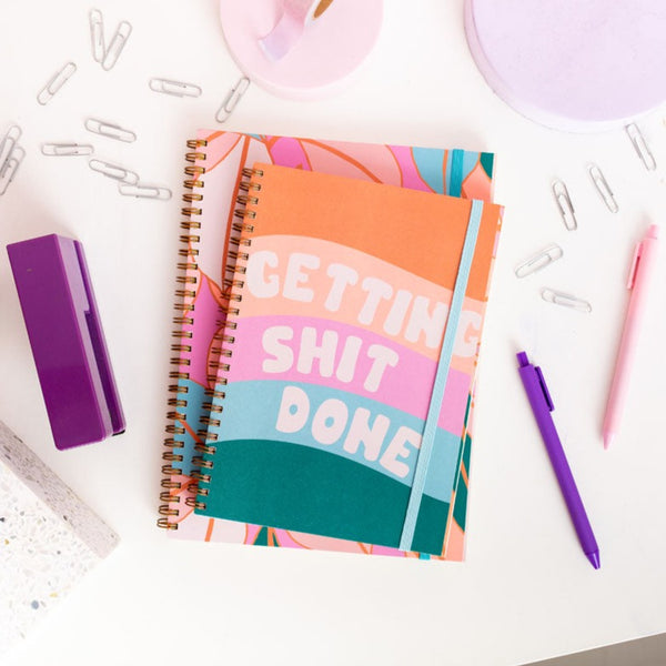 small goal getter planner with five waves in green, blue, pink, peach and orange, the words 'getting shit done' in white fluffy text at the front. surrounded by paper clips, two pen jotters, purple stapler and a lavender lamp.
