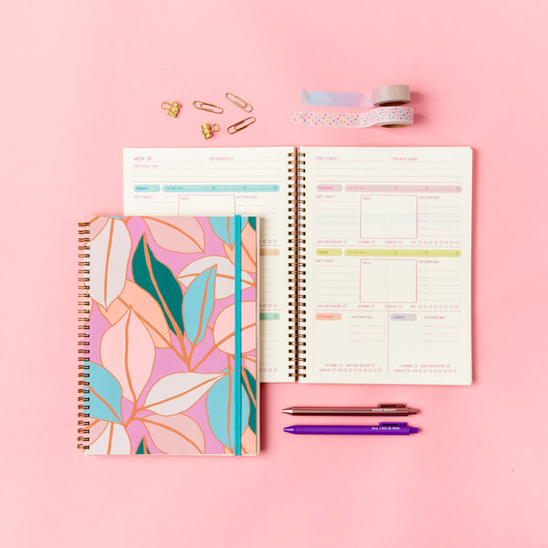Perpetual Planner - Goal Getter Lite - Talking Out Of Turn undated planner with pastel colored leaves in blue, teal, white, and pink on the cover. The opened planner shows the weekly undated to-do-list and reminders. Paperclips, wash-tape, and jotter pens around the planner on a pink background.