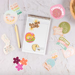 A task pad with multicolored and varying designed stickers and a pen scattered around. Sticker phrases are "Damn Girl," "Getting Shit Done," "Happy," "Be Kind,"" Manifest that shit," "Exhale the BS," "Chill the Fuck Out," and "Sorry our zodiacs just aren't compatible." Other stickers are arch shaped with either the moon, the sky, or both printed on in multiple neutral colors. Last sticker is of two abstract faces with thin line designs.
