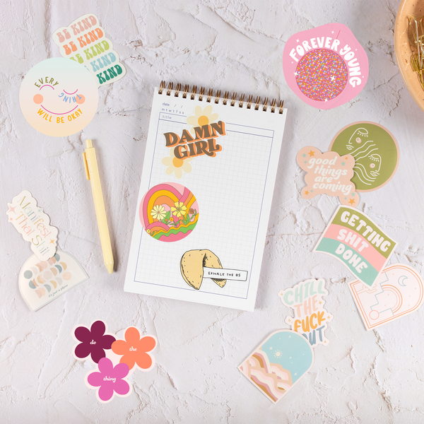 A task pad with multicolored and varying designed stickers and a pen scattered around. Sticker phrases are "Damn Girl," "Getting Shit Done," "Happy," "Be Kind,"" Manifest that shit," "Exhale the BS," "Chill the Fuck Out," and "Sorry our zodiacs just aren't compatible." Other stickers are arch shaped with either the moon, the sky, or both printed on in multiple neutral colors. Last sticker is of two abstract faces with thin line designs.