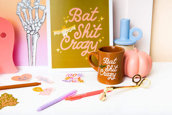 Camel color diner mug with "Bat Shit Crazy" written on the front in pink font, with illustration of bat and olive green minimalist sparkle-stars surrounding text.