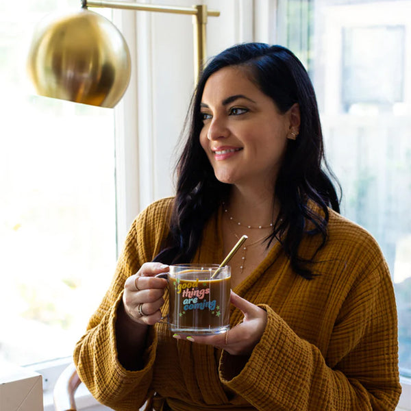 Woman in robe holding a clear glass mug with the phrase "good things are coming" printed on.