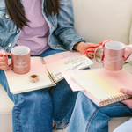 two women sitting writing notes in notebooks while holding metallic cup of ambition and reading is fundamental cups. 