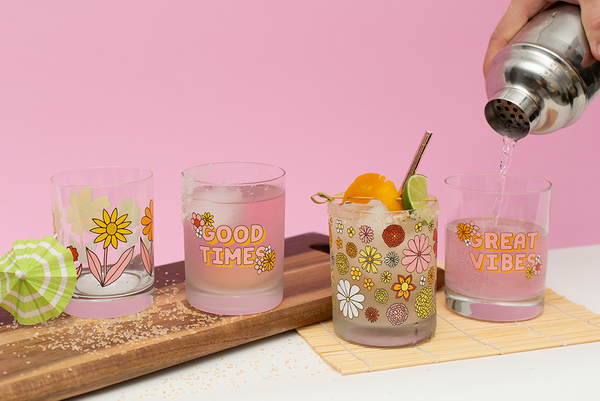person pouring water into a set of 4 retro flower power rocks clear glass set sitting on a white table with a pink wall in the background.