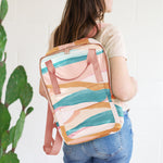 Vegan leather backpack with peach webbing and landscape pattern. Displayed by person holding back pack over their shoulder in front of white background.