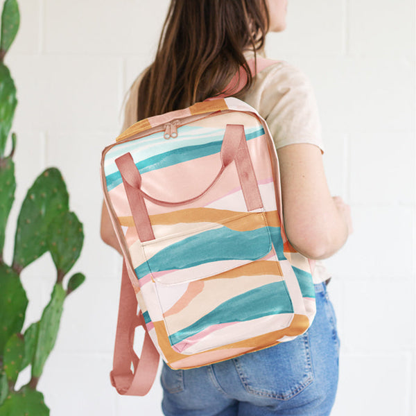 Vegan leather backpack with peach webbing and landscape pattern. Displayed by person holding back pack over their shoulder in front of white background.