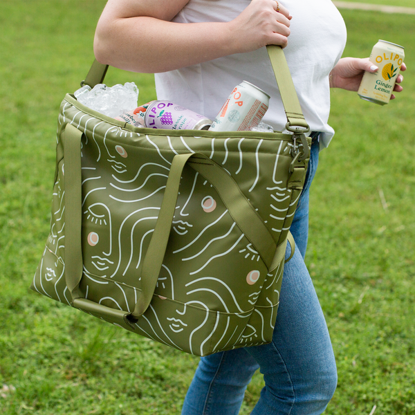 woman in blue jeans with white shirt carrying Ice Queen Zen Ladies shoulder cooler tote with olive green background and white retro ladies stencil drawing with pink and white cheeks.