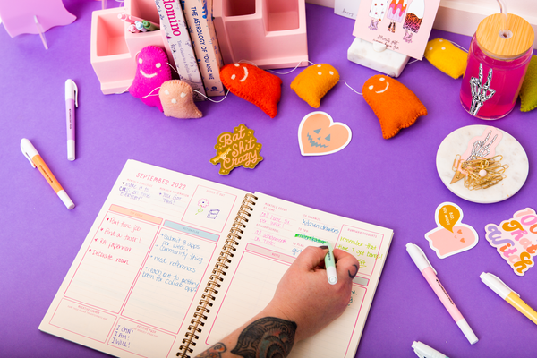 6 pastel highlighters displayed with TOOT Halloween Stickers and an Academic Planner. At the top of the picture is a Felt Multicolored Ghost Garland. There is also a Halloween Can Glass designed with a skeleton peace sign.