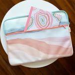 A laptop sleeve with sunset stripes across it sitting on a table with a notebook with the same design peaking out.